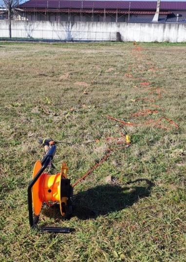 Stendimento di geofoni e bobina di cavo arancione su un prato durante un'indagine sismica MASW